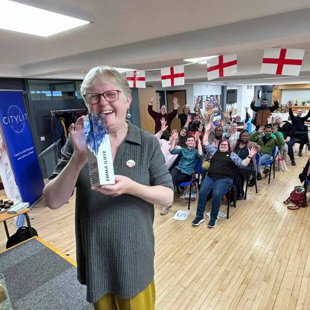 Emma, a smiling woman with short, light hair, holds an award with her name 'Emma Iliffe' on it. She stands in the foreground of a conference room. In the background, a group of people attending the conference are seated and enthusiastically waving their hands. The room is decorated with several St. George's flags hanging from the ceiling, and a banner on the left reads 'City Lit.' The atmosphere is joyful and celebratory.